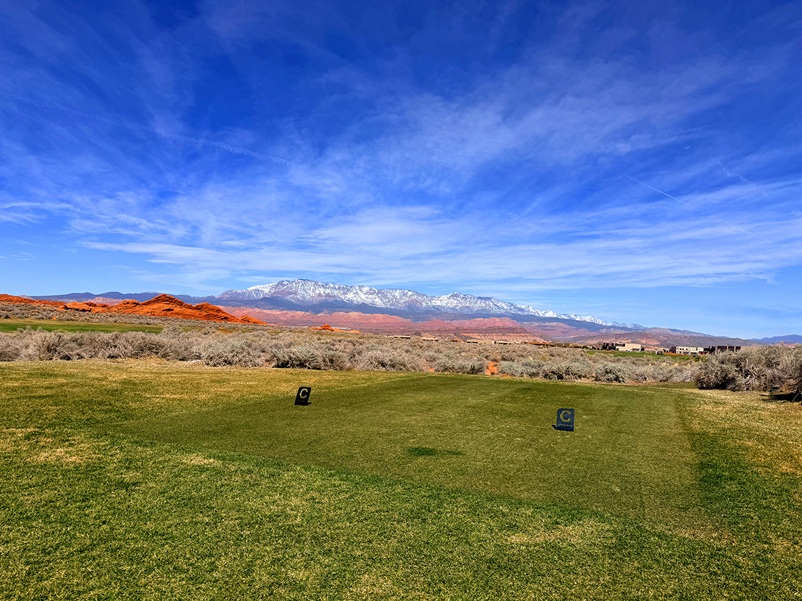 Wide view of a golf range with green grass, two markers, desert shrubs, orange hills, and snow-capped mountains under a blue sky.
